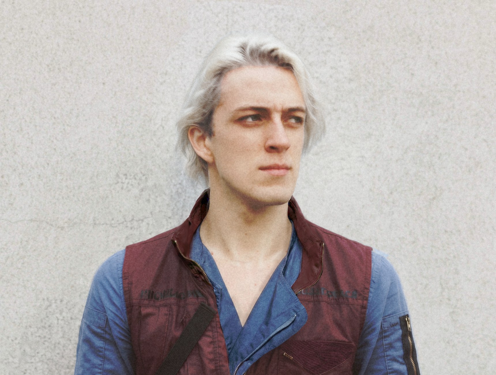 Jak Vanson against a pale wall, white-blond hair, burgundy vest over a blue shirt